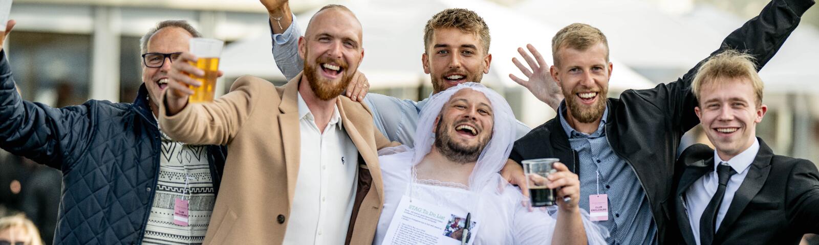 A stag dressed up in a veil and wedding dress, posing with his stag party at Windsor Races
