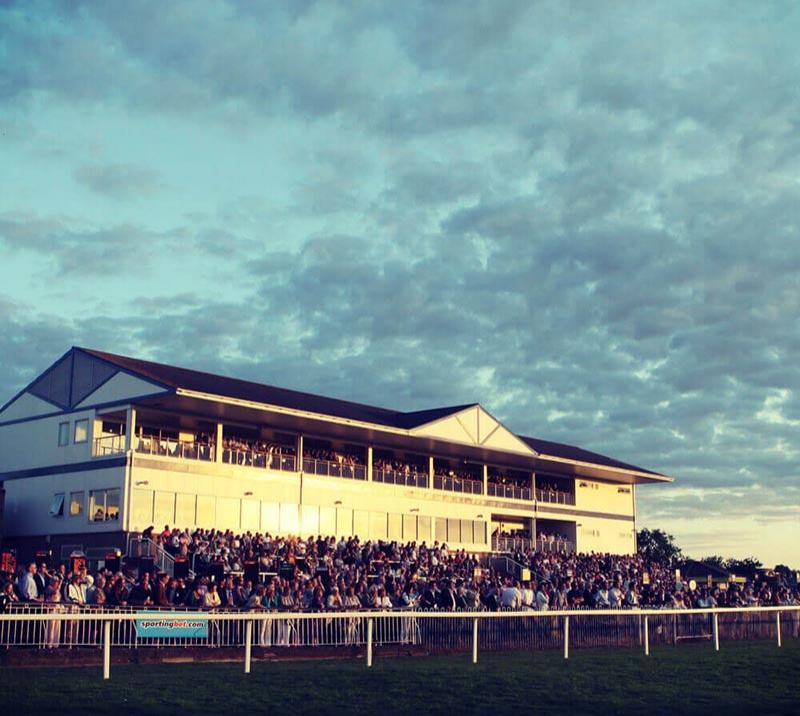 Evening race meeting at Windsor Racecourse, with crowds watching the action from the grandstand.